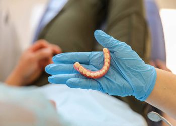 Close-up image of a dental professional wearing blue medical gloves and holding a lower denture. The photo was taken in a clinical setting and represents modern dental care, prosthetics, and oral health treatment.
