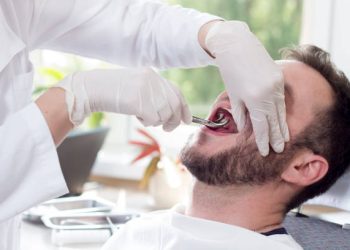 Tooth Extraction 6 A white man is sitting on a dental chair with his mouth wide open. The hands of the dentist in white gloves grip the tooth with claws.
